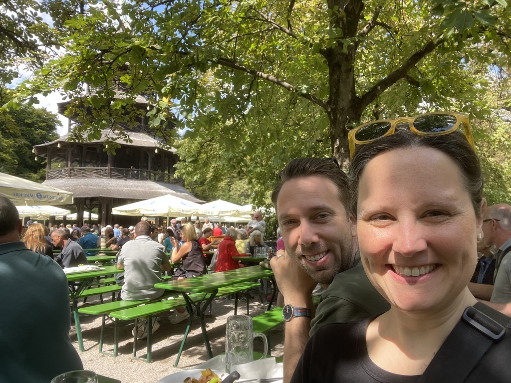 Spenser and Sarah enjoying a meal at the Biergarten in Englischer Garten, Munich, with a traditional wooden structure in the background and people dining at green picnic tables under sunny weather.
