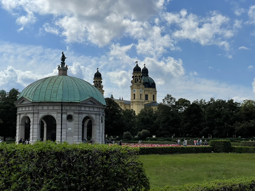 A view of a gazebo with a green dome in a park, surrounded by trees and people walking, with a large church tower in the background under a blue sky with clouds.