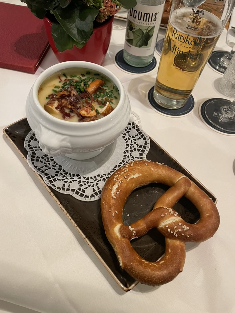 A bowl of potato soup topped with crispy onions and pretzel croutons and served with a pretzel on a plate, accompanied by a bottle of sparkling water and a glass of beer.
