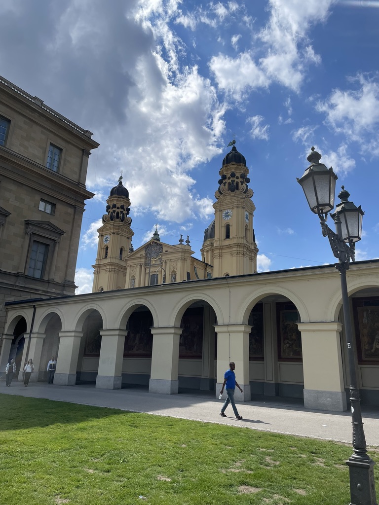 View of a building in Munich with its distinctive towers, flanked by columns and arches, under a partly cloudy sky.