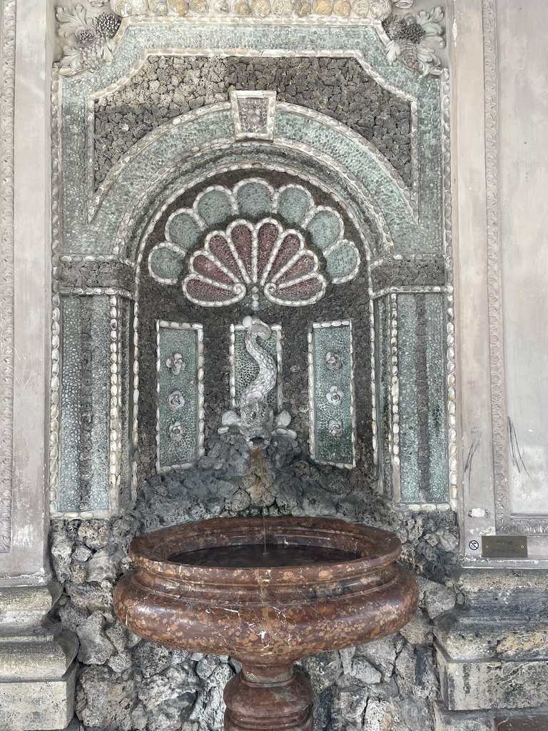 A decorative wall fountain featuring an ornate mosaic design with a central shell motif, flanked by intricate patterns and a dragon sculpture spouting water into a round basin.